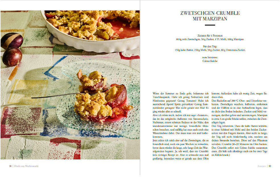 Aufgeschlagenes Kochbuch »Frisch vom Wochenmarkt« mit Rezept für Zwetschgen Crumble mit Marzipan. Eine Portion des Crumbles ist auf einem Teller angerichtet.