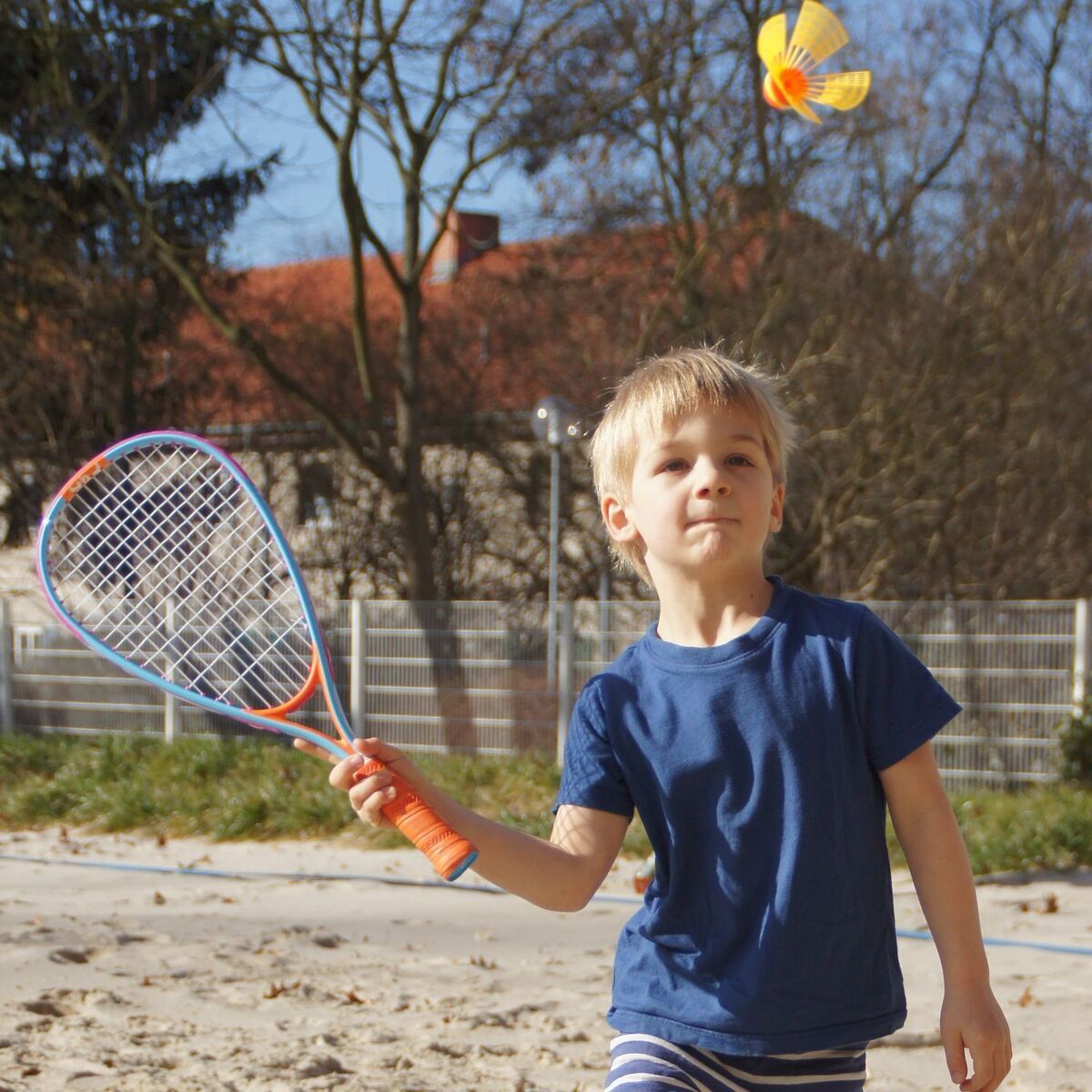 Ein Kind spielt im Freien mit dem Speedminton »Fun-Set«, das aus einem Schläger und einem speziellen Federball besteht. Es trägt ein blaues T-Shirt und blickt auf den fliegenden Federball. Bäume und ein Haus sind im Hintergrund zu sehen.