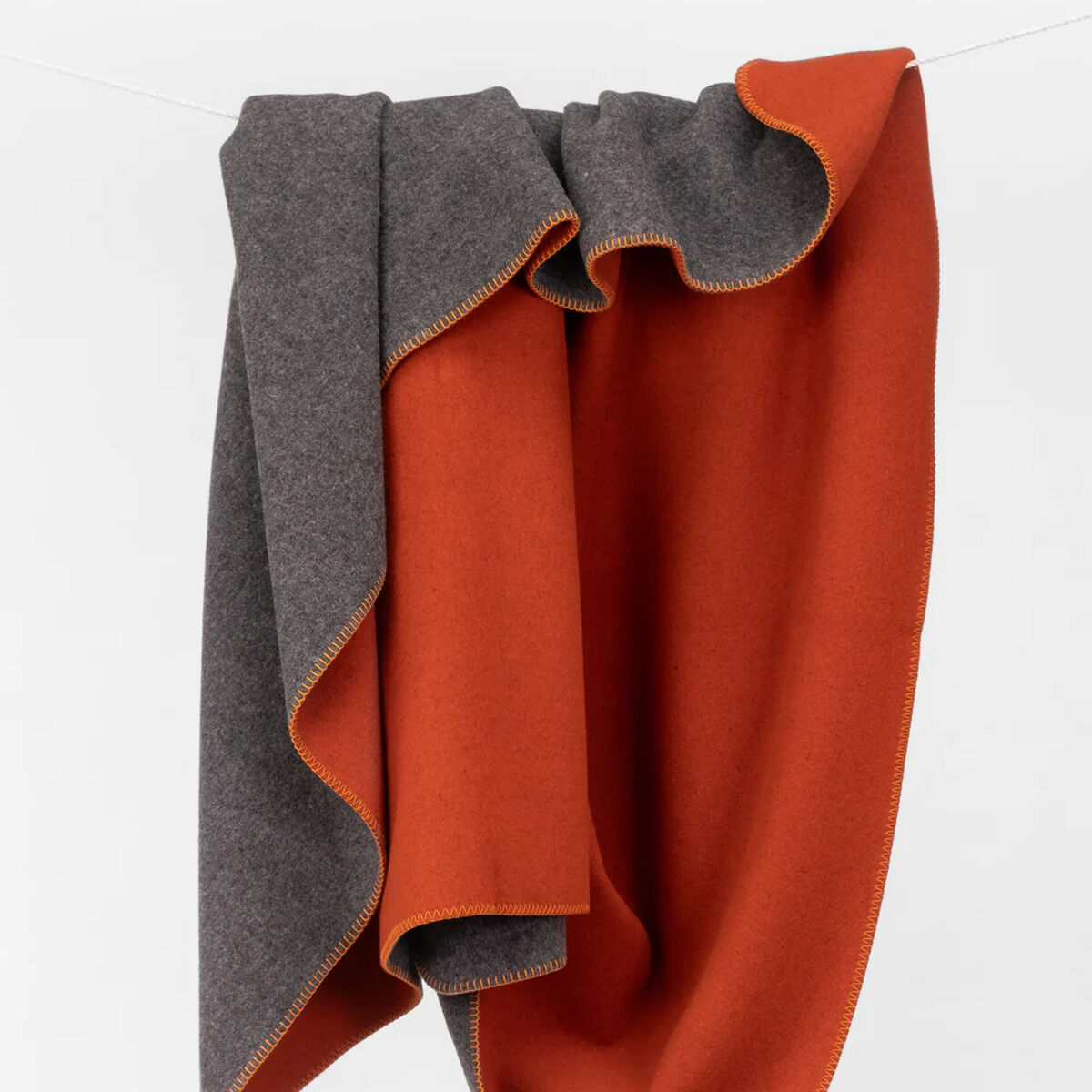 Die Merinodecke »FireSideMoon« von COASTLAND hängt locker drapiert. Die Decke ist außen glutrot und innen anthrazit mit gelber Kettelnaht, aus feiner, unbehandelter Merinowolle gefertigt und wirkt weich, hochwertig und flauschig – ideal für gemütliche Stunden zuhause.