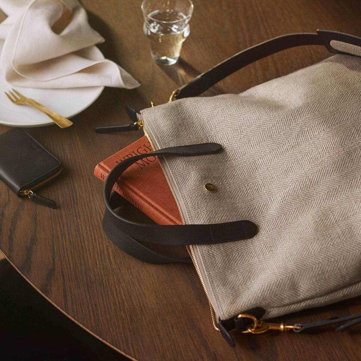 Eine beige MISMO-Shopper-Tasche mit schwarzen Lederriemen liegt auf einem Holztisch neben einem weißen Teller mit goldener Gabel, einem kleinen schwarzen Geldbeutel und einem Glas Wasser.