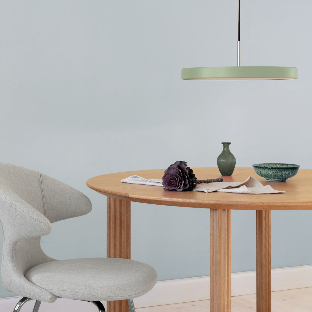 Pendelleuchte »Asteria Medium« über einem runden Holztisch mit Dekoration in einem modernen und minimalistischen Raum. Die Wände sind hellblau, der Stuhl ist hellgrau und auf dem Tisch liegen einige Objekte.