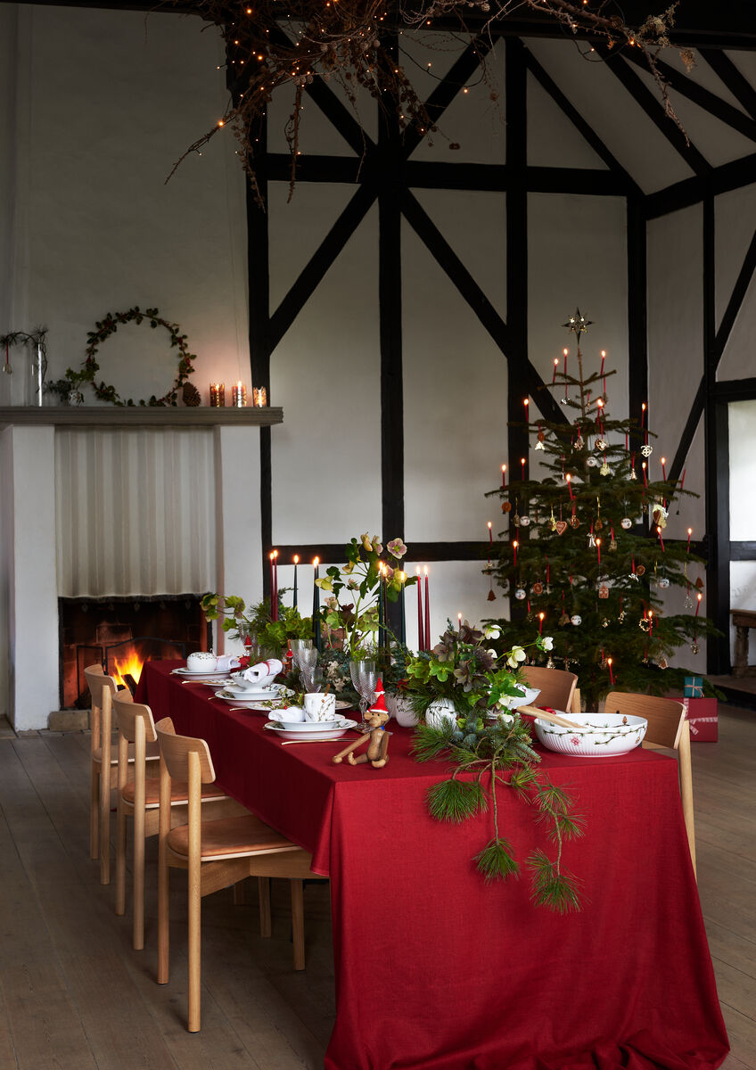 Ein festlich gedeckter Tisch in einem Esszimmer, geschmückt mit rotem Tischtuch und Weihnachtsdekoration. Im Hintergrund steht ein beleuchteter Weihnachtsbaum nahe einem Kamin. Eine Weihnachtsschale der Marke »Hammershøi« ist auf dem Tisch arrangiert. Der Raum ist in einem modernen, skandinavischen Stil eingerichtet.