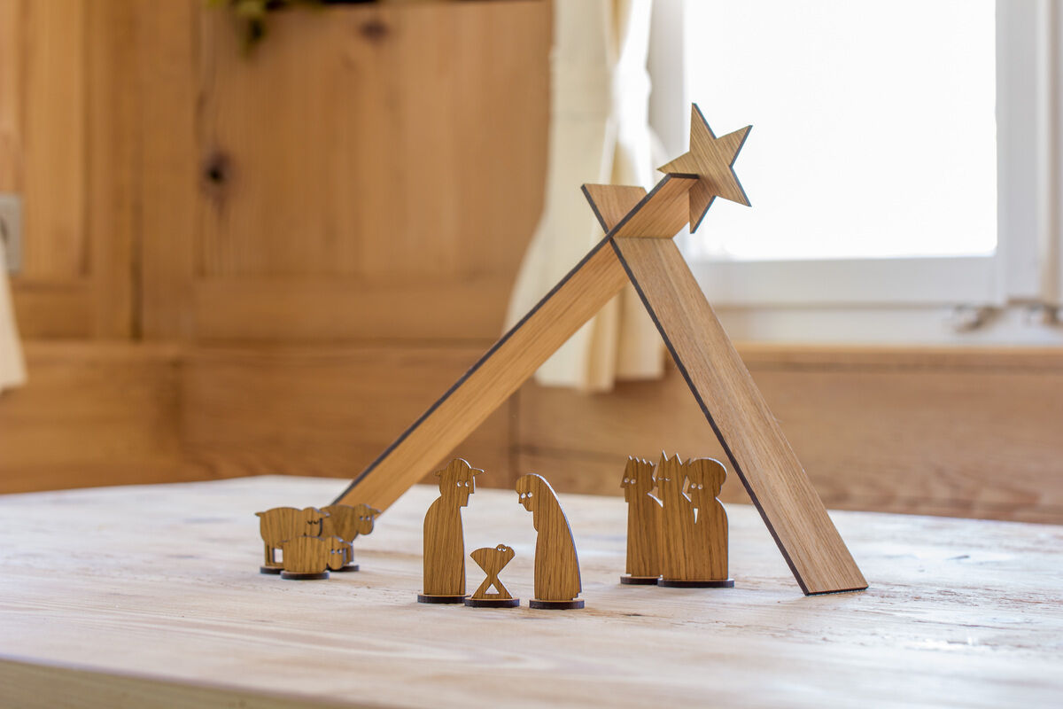 Eine minimalistische Weihnachtskrippe aus Holz steht auf einem Tisch. Die Szenerie zeigt Figuren von Maria, Josef, dem Jesuskind, einem Schäfer mit seinen Schafen und einem Stall mit einem Stern oben drauf. Es handelt sich um eine handgefertigte und einfache Darstellung der Geburt Jesu.