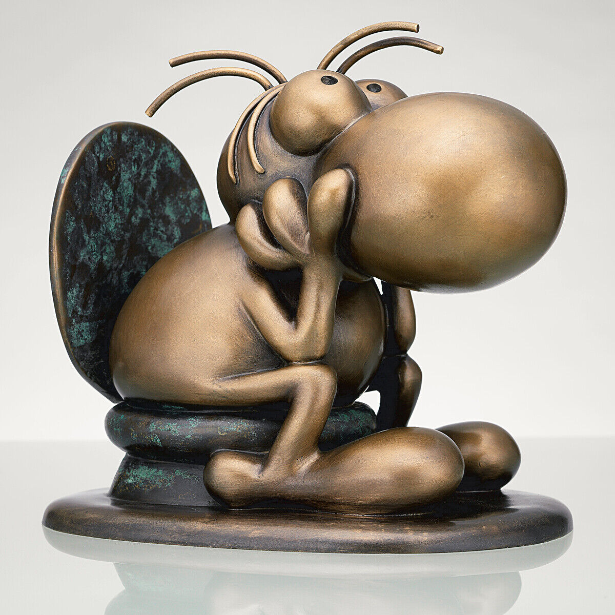 Bronzeskulptur einer cartoonhaften Figur im Stil von Mordillo, die nachdenklich auf einem Podest sitzt. Die Skulptur zeigt eine humorvolle, stilisierte Darstellung in Bronze- und Grün-Tönen.