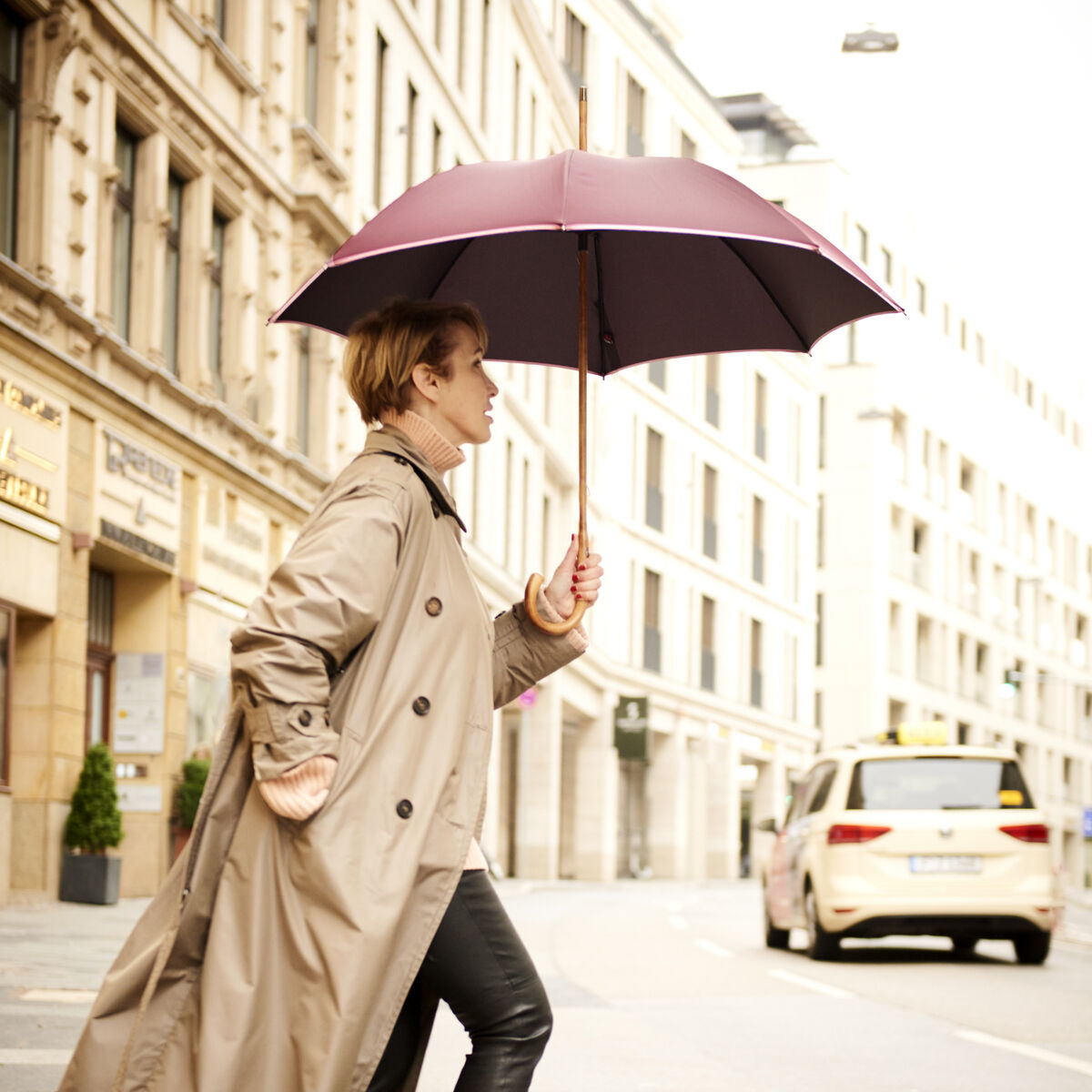 Eine Frau mit einem beerenfarbenen Regenschirm und hellem Trenchcoat überquert an einem Regentag eine städtische Straße. Im Hintergrund sind Gebäude und ein Auto zu sehen.