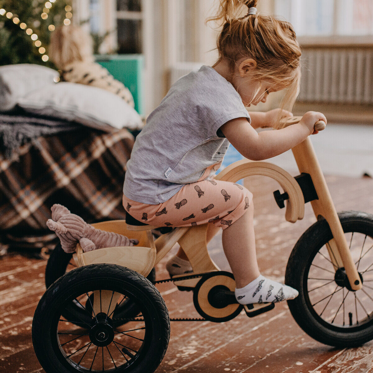 Ein kleines Kind fährt drinnen auf einem Dreirad aus Holz mit schwarzer Bereifung und Erweiterung für das Balance Bike. Im Hintergrund sind ein Sofa mit einer karierten Decke, ein Weihnachtsbaum und eine gemütliche Raumatmosphäre zu sehen. Das Kind trägt helle Kleidung mit Mustern.