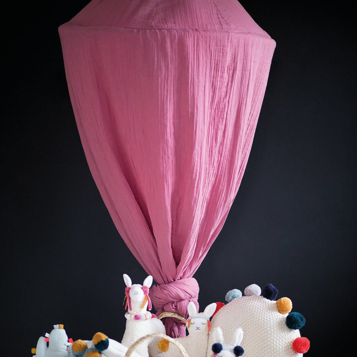 Ein rosa Kinder-Betthimmel aus Baumwollematerial, aufgehängt über einem Bett oder einer Kuschelecke. Im Vordergrund sind weiße und bunte Dekorationselemente wie Kissen oder Kuscheltiere zu sehen.