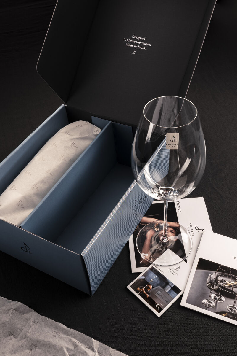Ein Rotwein 2er-Set »Enoteca« bestehend aus einem eleganten Weinglas und einer stilvollen Geschenkverpackung. Das Set liegt auf einem dunkelgrauen Hintergrund mit einigen Prospekten daneben.