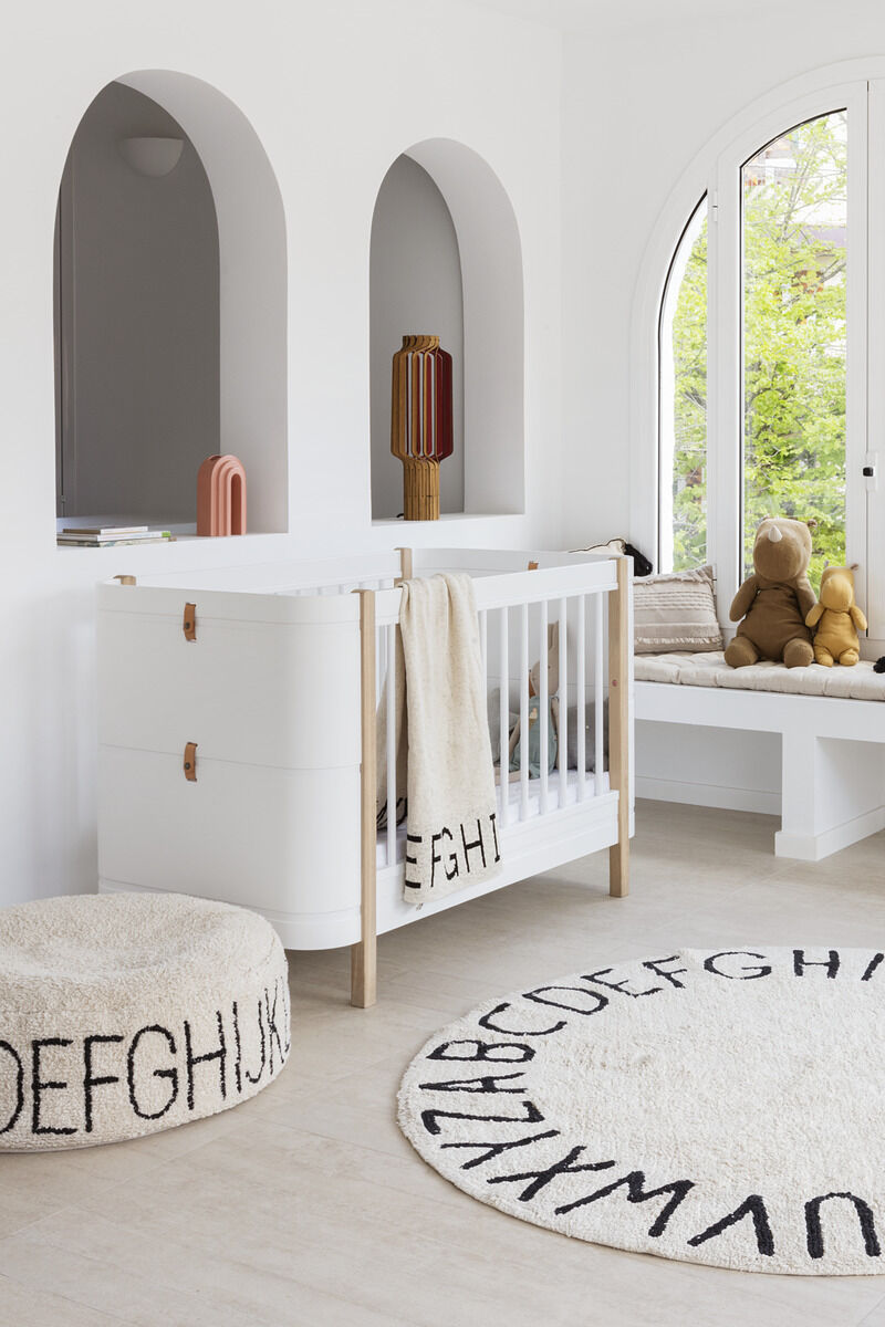 Ein helles und freundliches Kinderzimmer mit einem Pouf mit Alphabet-Aufdruck, einem passenden Teppich und einem weißen Gitterbett. Das Zimmer ist minimalistisch gestaltet und mit Spielzeug dekoriert.