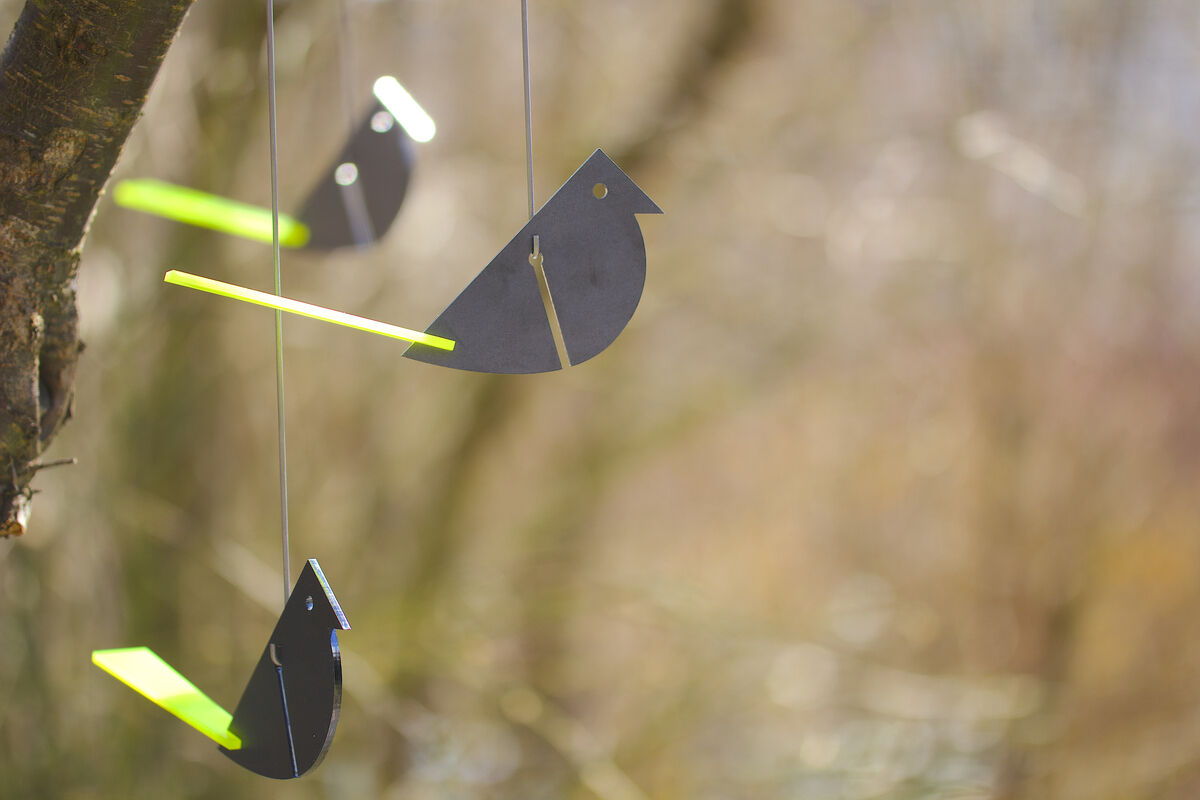 Holzvögel in Form von »Anton Grünfink« hängen an Fäden an einem Baumzweig. Einige Teile sind in hellem Grün und Schwarz bemalt. Im Hintergrund unscharf, Naturkulisse in Braun- und Grüntönen.