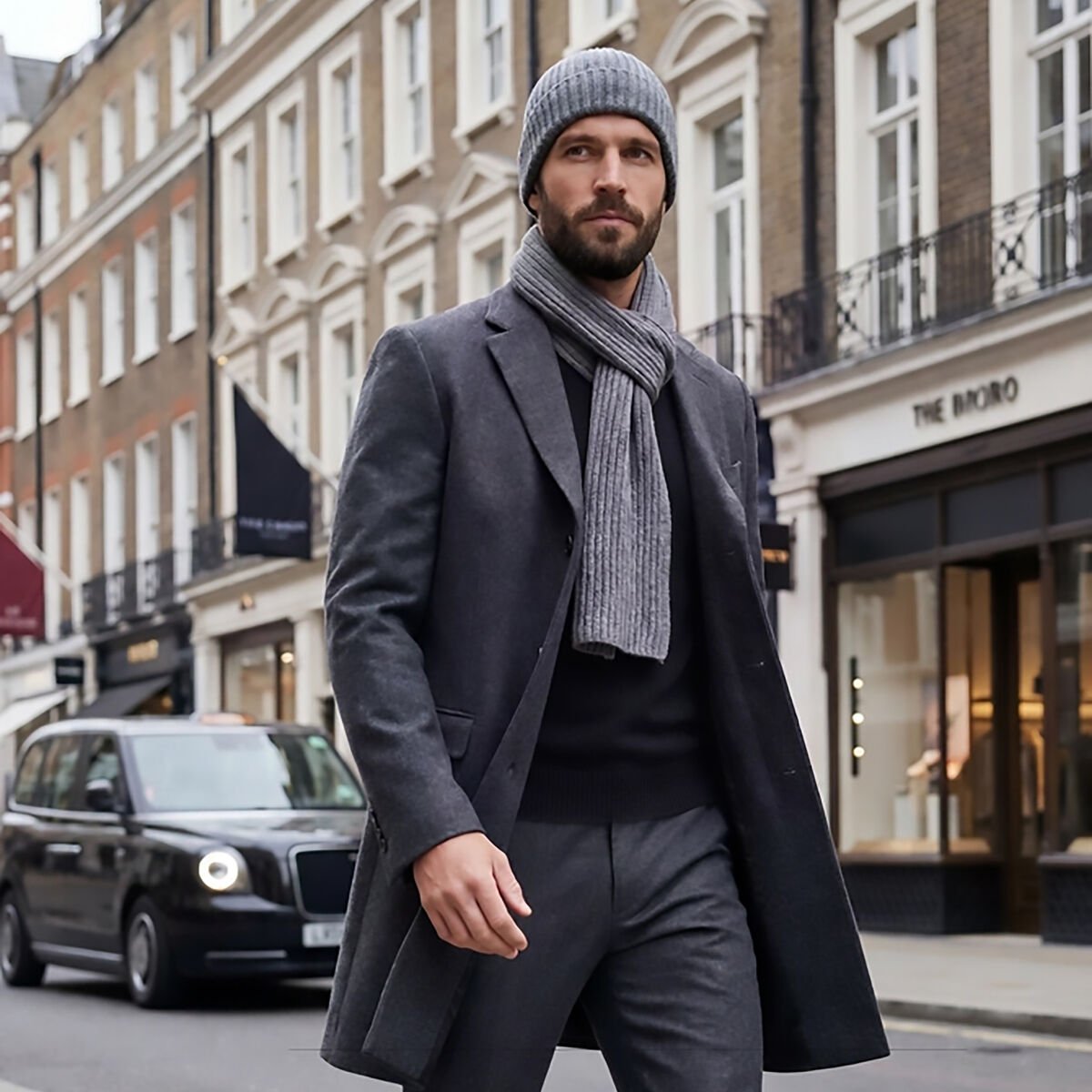 Ein Mann geht in einer Stadtstraße und trägt einen eleganten grauen Mantel, einen graumelierten Kaschmir-Schal "Dominik" um den Hals, dazu graue Hose, schwarzen Pullover, graue Strickmütze und braune Lederschuhe. Im Hintergrund klassische Stadthäuser und ein schwarzes Taxi.