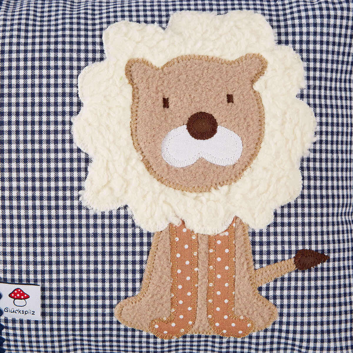 Ein handgemachtes Namenskissen mit einem applizierten Löwenmotiv auf einem blauen und weißen Gittermusterstoff. Der Löwe hat braune Filzohren und Pfoten, eine beige Mähne und einen rosa-weiß gepunkteten Körper.