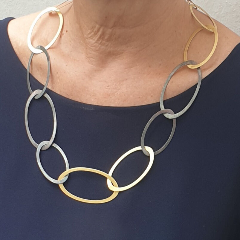Das Collier »Silberringe« von Maria Rzewuska ist ein elegantes Schmuckstück aus teils oxidiertem, teils feingoldplattiertem 925er Silber. Große, ineinandergreifende, ovale Ringe schmücken ein dunkles Oberteil. Das Design wirkt modern und vielseitig tragbar.