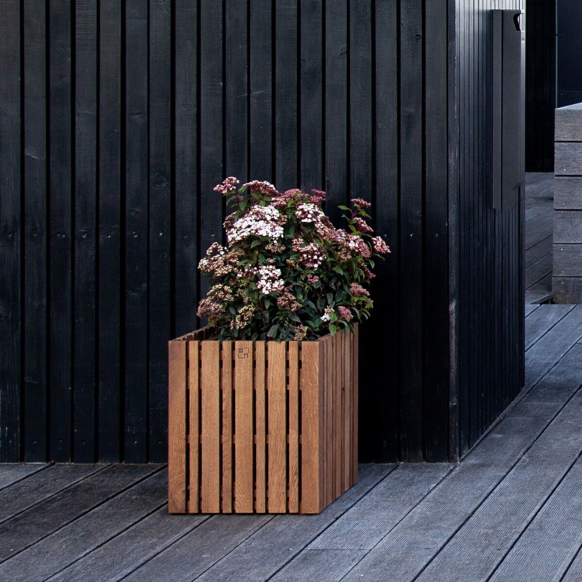 Pflanzgefäß »GrowMORE« in Holzoptik auf einer Terrasse vor einer modernen schwarzen Wand. Das Gefäß enthält blühende Pflanzen mit grünen Blättern und rosa-weißen Blüten.