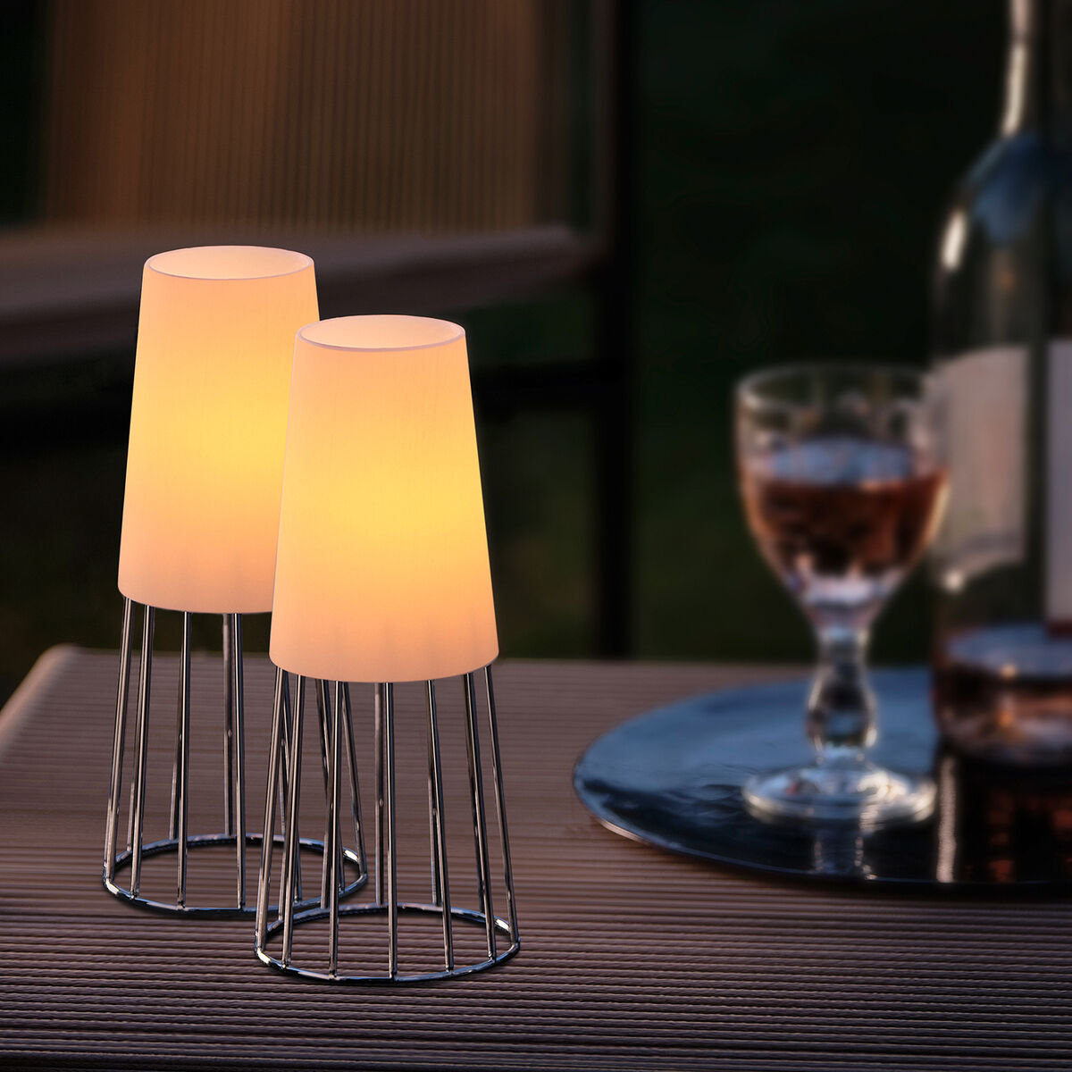 Zwei Teelichter aus der Serie 'Lighthouse' stehen auf einem Tisch im Freien, in der Dämmerung. Die Teelichter haben eine moderne, minimalistische Ästhetik mit weißen Leuchten und einem schwarzen Metallrahmen. Im Hintergrund ist ein Glas mit Roséwein und eine Flasche auf einem Tablett zu sehen.