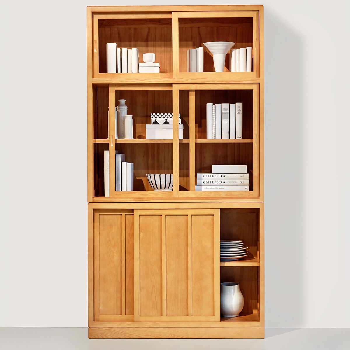 Die Vitrine »Multimodus« von MARKTEX zeigt eine hohe, zweigeteilte Vitrine aus honigfarben lasiertem Massivholz mit sechs Schiebetüren. Im oberen Teil sind klare Glastüren eingesetzt, dahinter Bücher und Dekorationsobjekte. Unten befinden sich mit Holzfront versehene Schiebtüren, teils offen, mit Tellern und Keramik dekoriert.