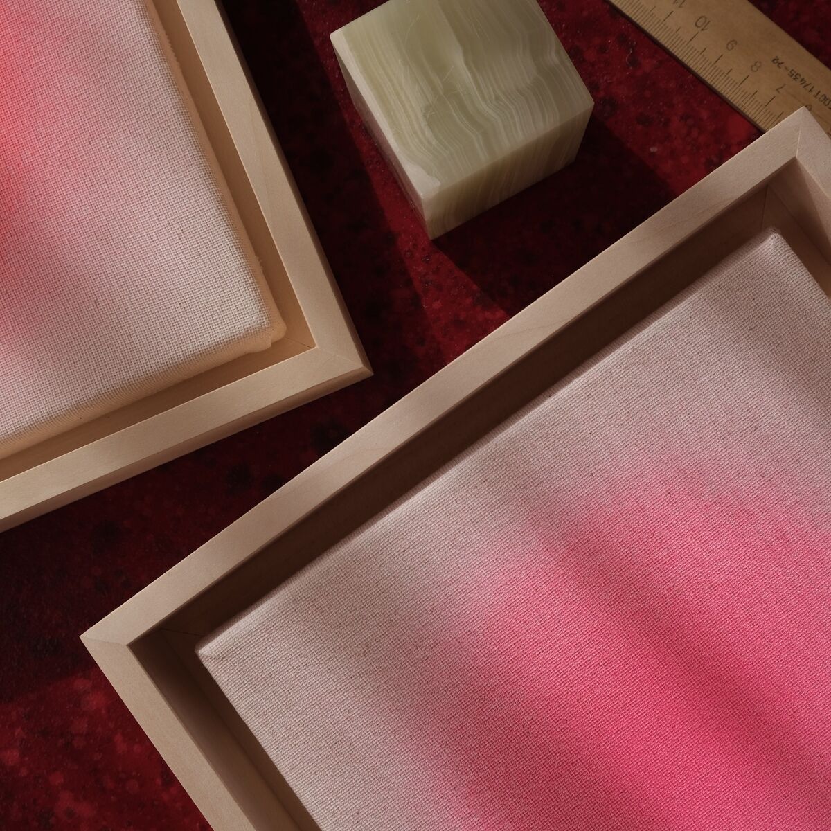 Zwei quadratische Bilder in Holzrahmen zeigen einen zarten Farbverlauf in Pink- und Rosatönen auf strukturiertem Untergrund. Die Werke liegen auf einem roten Untergrund, daneben befinden sich ein Lineal und ein grünlicher Würfel. Das Bild vermittelt einen modernen, minimalistischen und künstlerischen Eindruck.