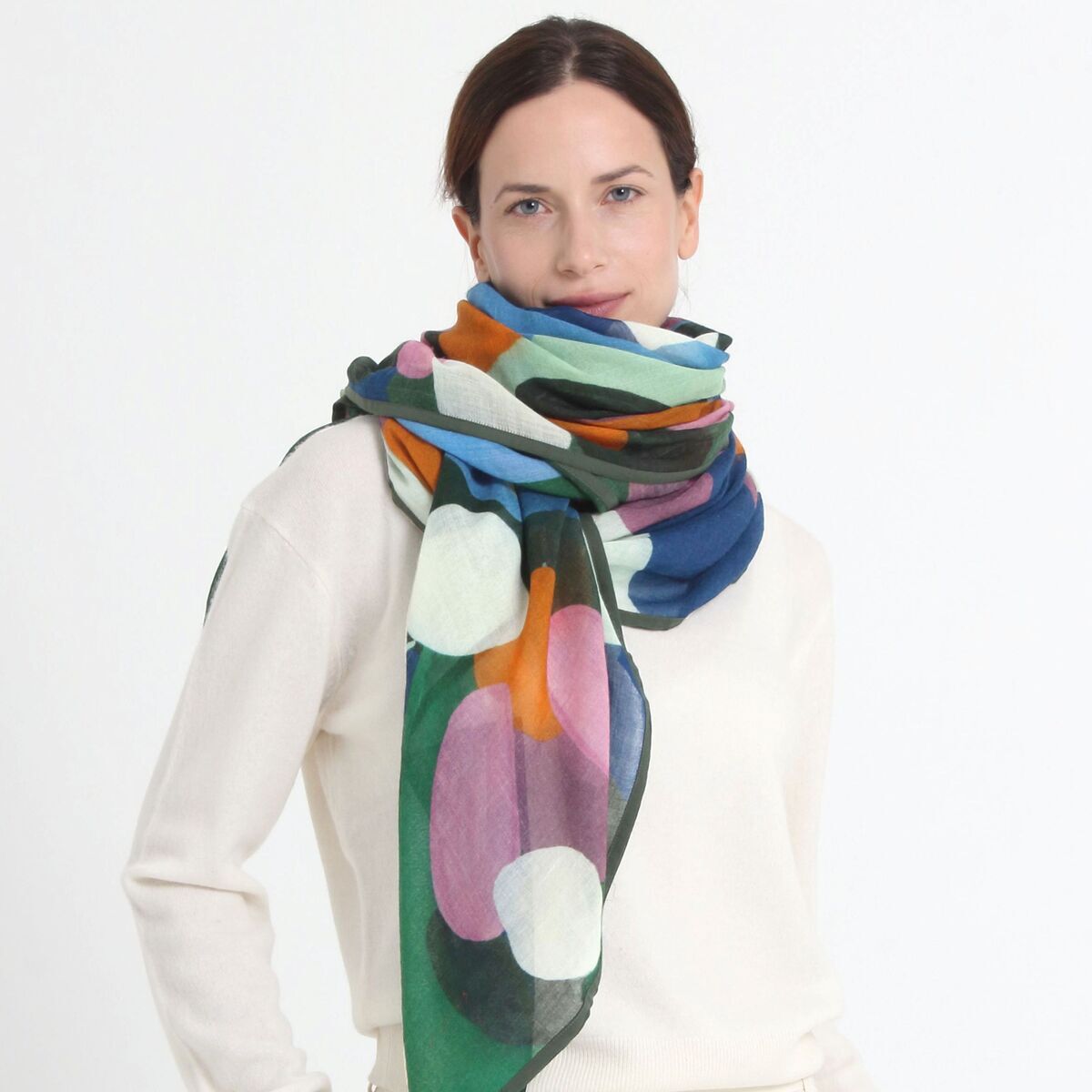 Eine Frau trägt einen weißen Pullover und präsentiert den Wollschal »Evie« Grün von Shalls Barcelona. Der Schurwollschal zeigt ein großes, abstraktes Muster in Grün, Blau, Orange, Weiß, Lila und Rosa. Das Gesamtbild wirkt modern, stilvoll und farbenfroh.