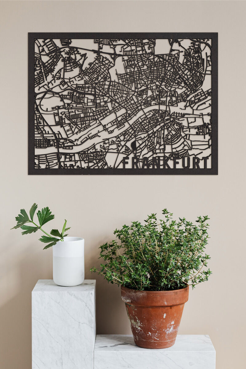 Der Stadtplan von Frankfurt aus Holz zeigt filigran die Straßen und Gebäude der Stadt. Das Kunstwerk hängt an einer beigen Wand über einem weißen, marmorähnlichen Sockel und einem Terrakottatopf mit einer grünen Pflanze.
