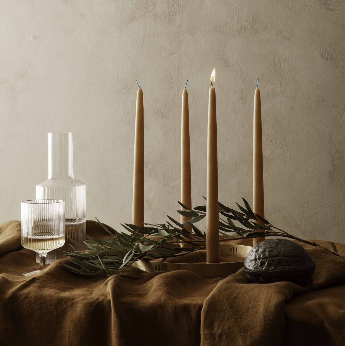 Stilvolle Tischdekoration mit einem minimalistischen Kerzenhalter 'Circle' und mehreren brennenden Stabkerzen. Der Tisch ist mit braunem Stoff bedeckt, umgeben von Olivenzweigen und verschiedenen Glasgefäßen.