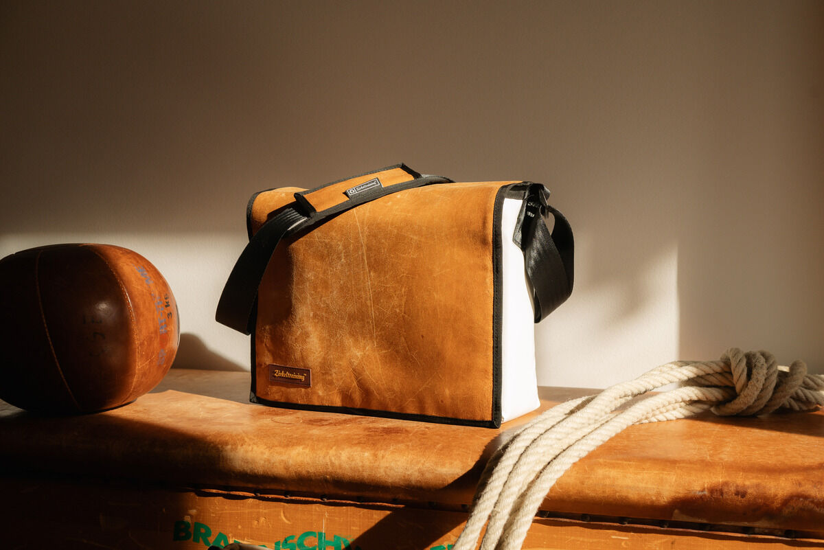 Eine ZEIT-Tasche »Kasten« mit einem schwarzen Gurt liegt auf einer braunen Oberfläche. Die Tasche besteht aus braunem Leder mit weißen Seiten, und ein schwarzer Gurt ist sichtbar. Im Hintergrund sind ein alter Lederball und ein Stück Seil zu sehen.