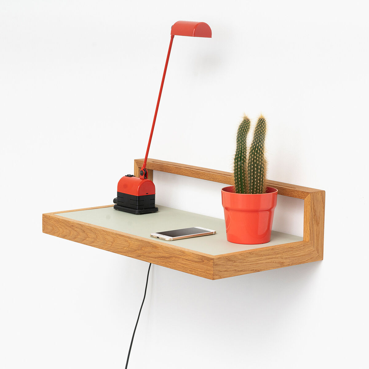 Ein modernes Wandboard aus Holz in der Farbe Pistazie, das mit einer roten Schreibtischlampe, einem roten Blumentopf mit einem kleinen Kaktus und einem Smartphone dekoriert ist. Das Wandboard hat ein minimalistisches Design und bietet praktischen Stauraum.