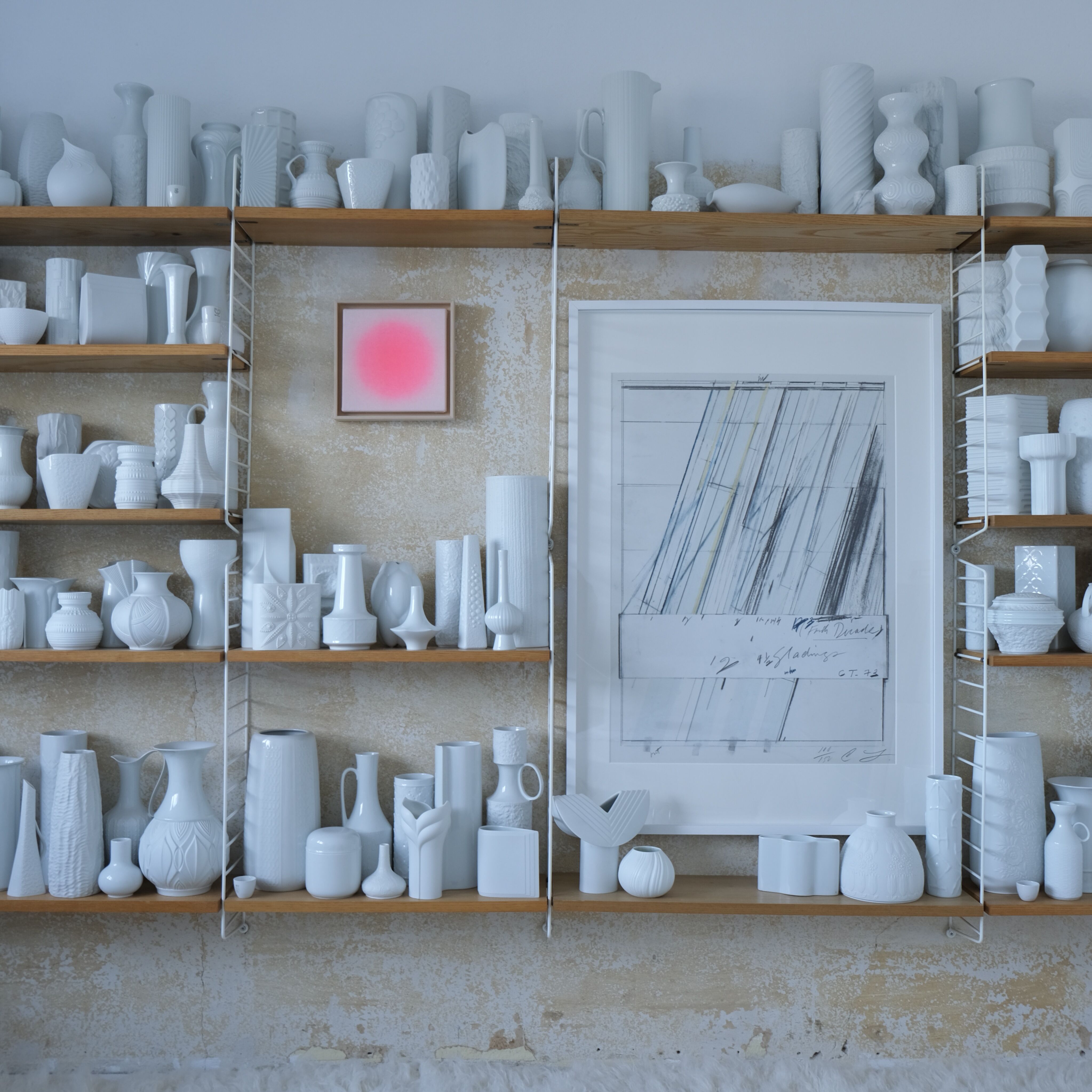 Großformatige moderne Kunstinstallation an einer Wand: Auf hellen Holzregalen stehen viele weiß glasierte Vasen und Gefäße in unterschiedlichen Formen. Zwischen den Regalen hängen zwei gerahmte Bilder. Das linke Bild zeigt ein leuchtend apricotfarbenes Quadrat, das rechte eine abstrakte schwarz-weiße Zeichnung. Die Wand dahinter ist beigefarben, teilweise alt und abgenutzt.