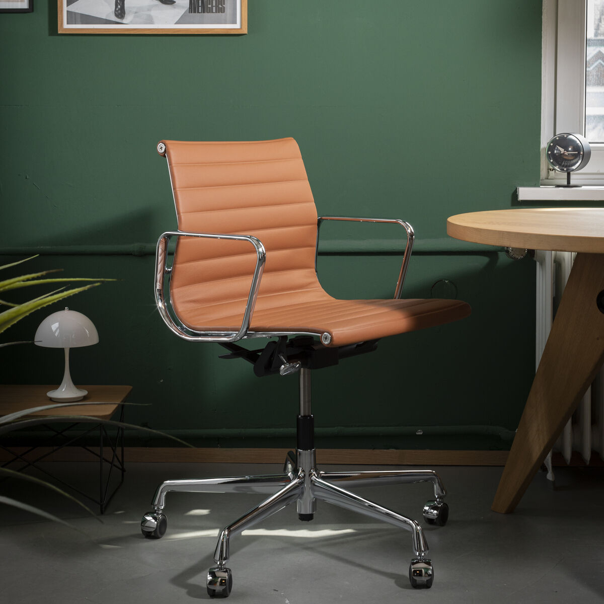 Ein moderner Bürostuhl, der Vitra Alu Chair EA 118 in verchromtem Design, steht vor einem grünen Wandanstrich. Der Stuhl hat eine gepolsterte braune Lederbespannung und drehbare Rollen. Neben dem Stuhl steht ein runder Holztisch, dokumentiert durch rahmenlose Fenster. An der Wand hängen mehrere Bilder, darunter eines mit dem Wort 'CANCELLED' und ein Schwarz-Weiß-Foto.