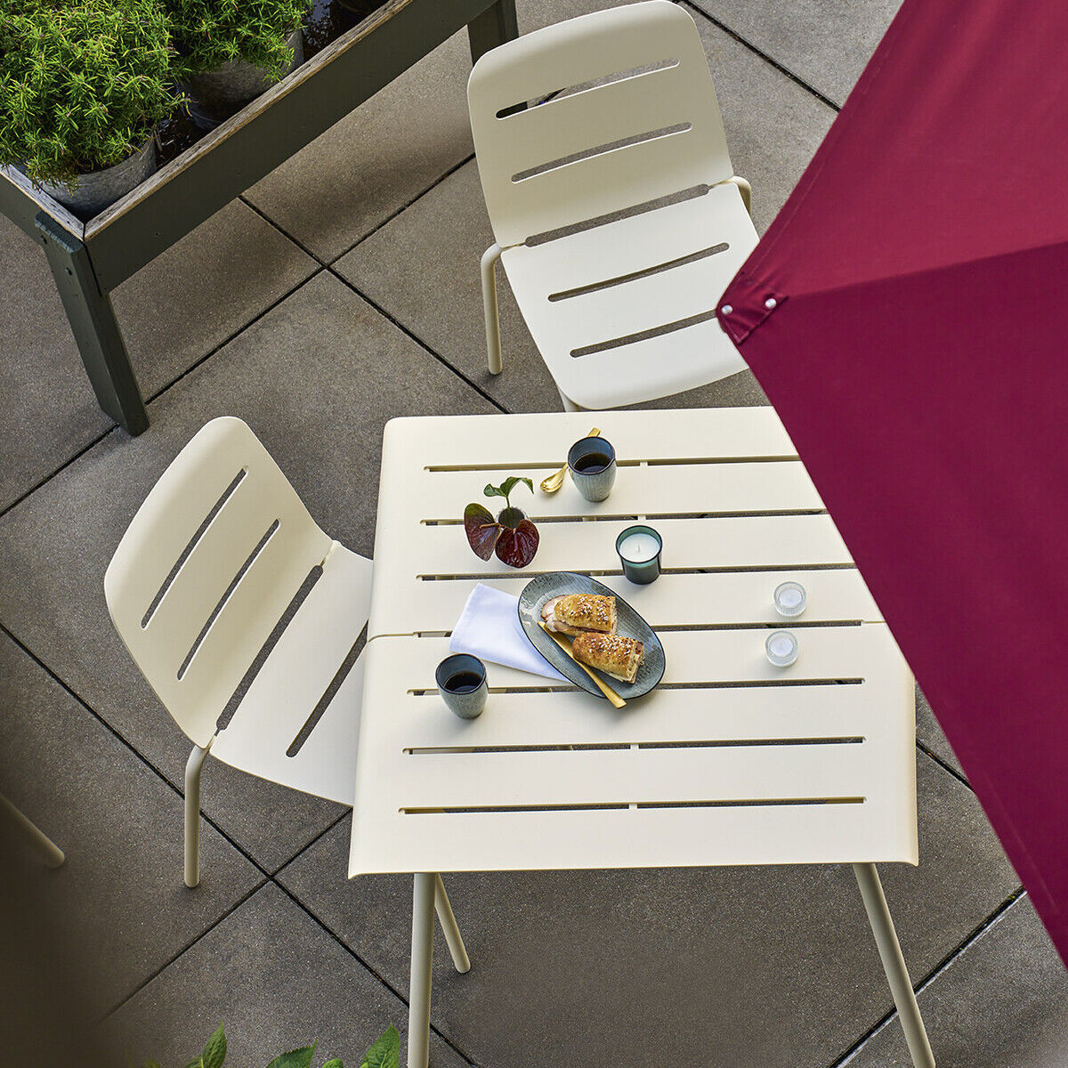 Ein moderner Terrassentisch, Modell »VAPIO«, 90x90 cm, beige, mit zwei passenden Stühlen, auf einer gefliesten Terrasse platziert. Ein roter Sonnenschirm spendet Schatten. Der Tisch ist teilweise gedeckt, mit Gläsern und einem kleinen Servierbrett. Im Hintergrund ist ein Teil eines grünen Pflanztrogs zu sehen.