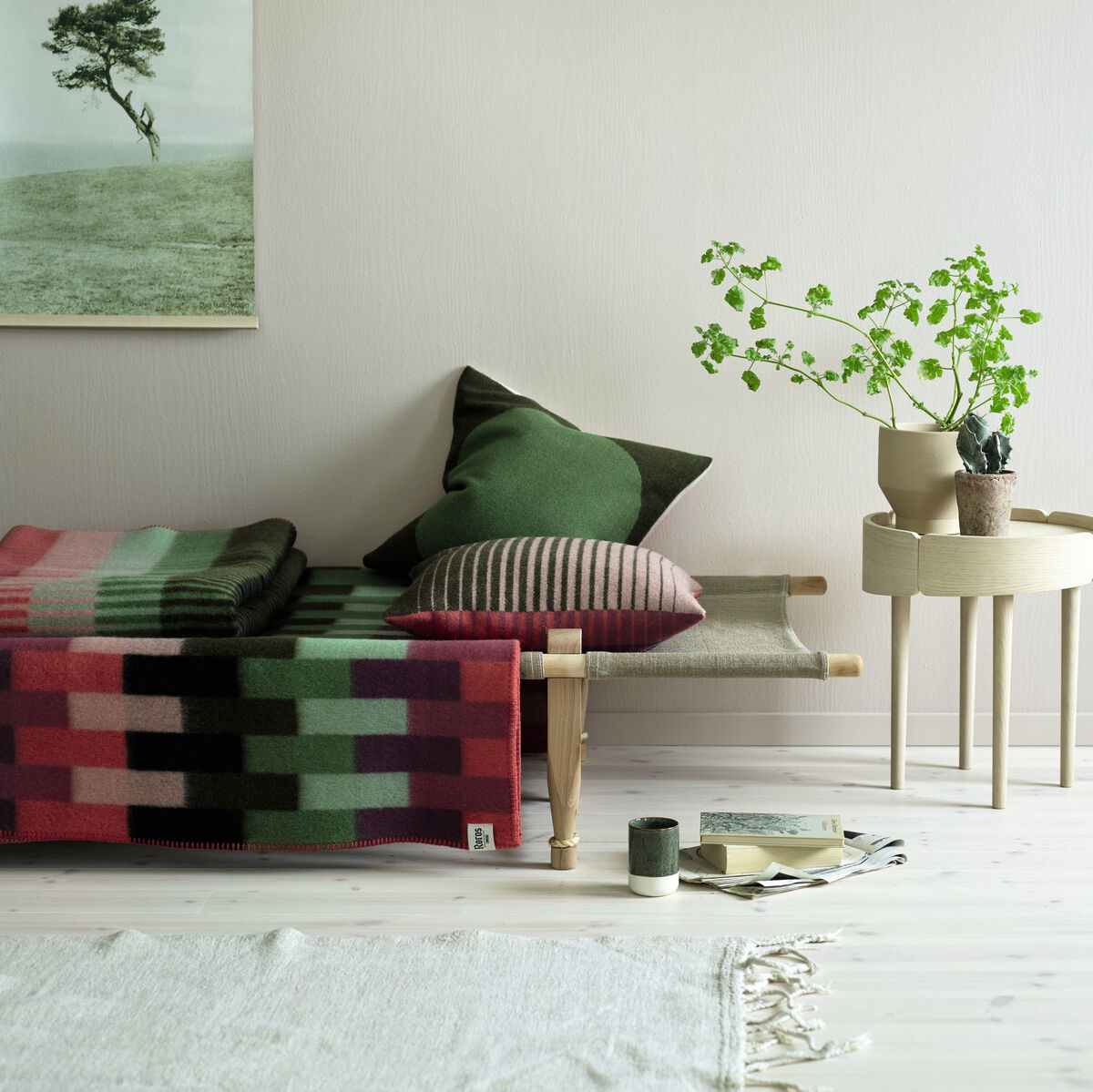 Ein stilvolles Wohnzimmer mit einer auf einem Sofa drapierten Lammwolldecke in mehreren Farben, darunter Mintgrün, Hellrosa und Dunkelgrün. Daneben befinden sich dekorative Kissen in verschiedenen Grüntönen und ein kleiner Beistelltisch mit einer Pflanze und einigen Büchern.