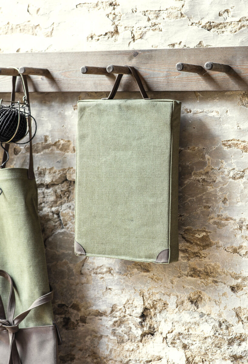Ein grünes Kissen aus Canvas-Material, das an einem rustikalen Holzhaken an der Wand hängt. Im Hintergrund ist eine raue Steinmauer sichtbar. Neben dem Kissen hängt ein grüner Gartenwerkzeug-Beutel.