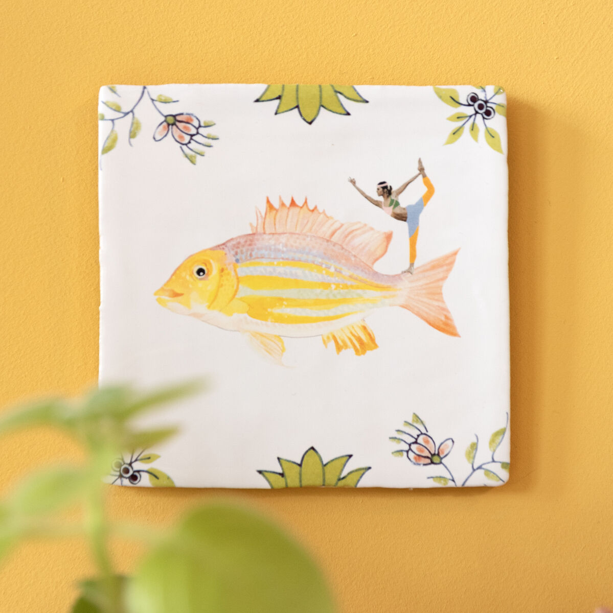Keramikfliese »Im Flow«: Eine handbemalte Fliese mit einem gelben Fischmotiv und dekorativen Blumen auf einem weißen Hintergrund, aufgehängt an einer gelben Wand. Im Vordergrund sind unscharfe grüne Blätter und ein Korallendekor zu sehen.