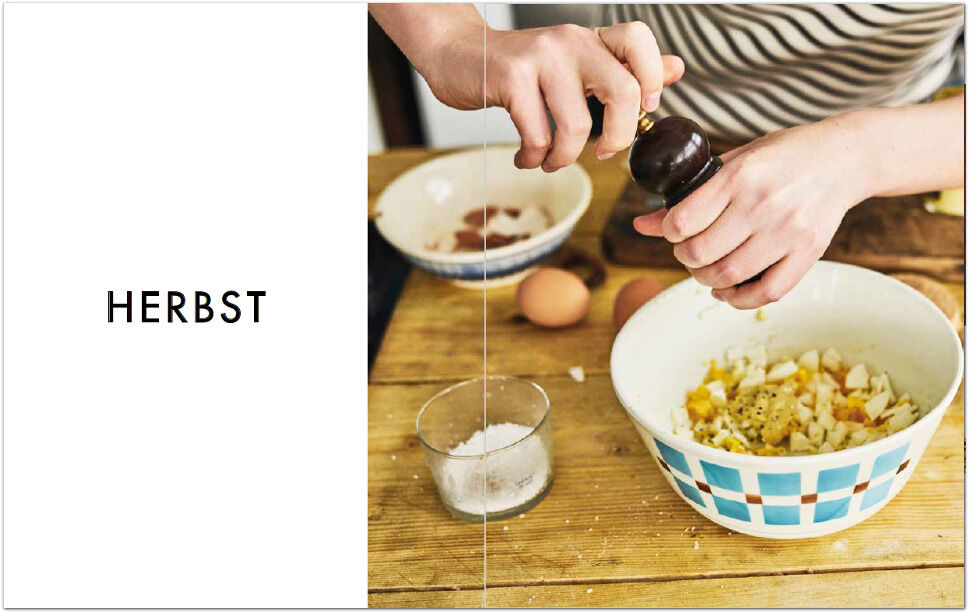 Hände einer Person, die Pfeffer in einen Salat aus einer großen Schüssel streut. Neben der Schüssel befinden sich weitere Zutaten wie Eier und Salz. Das Bild ist in einem Kochbuch mit dem Titel 'Herbst'.