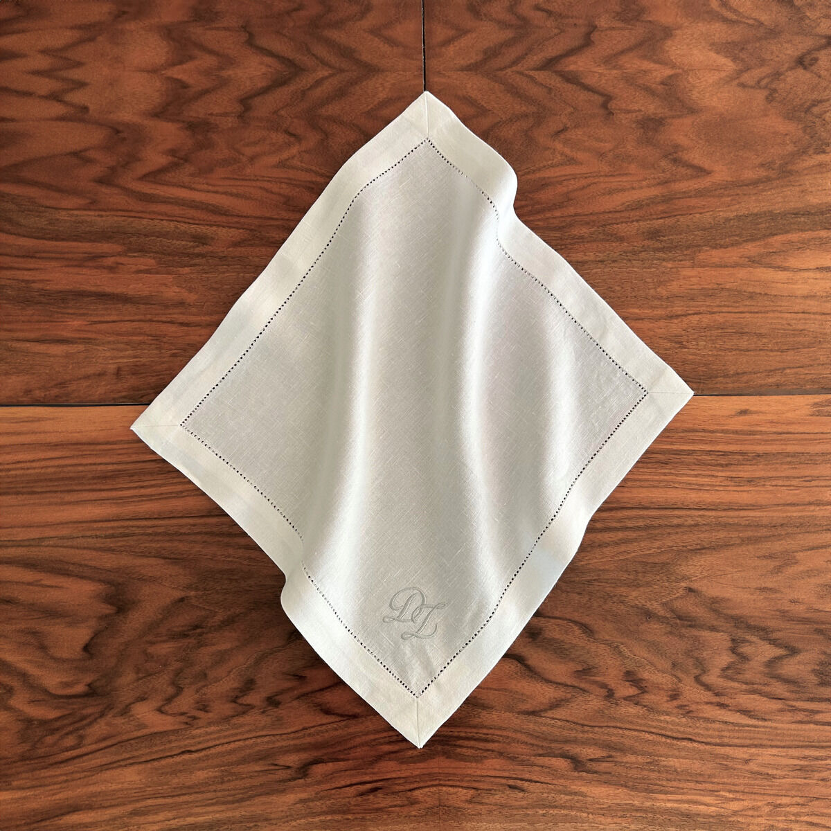 Eine weiße Leinenserviette mit Monogramm auf einem Holztisch. Die Serviette ist elegant bestickt und kunstvoll drapiert.
