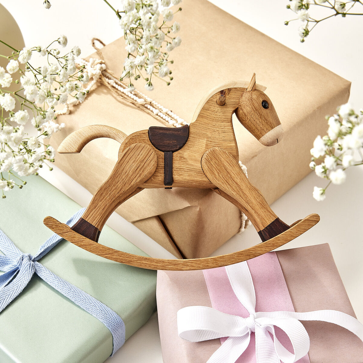 Auf dem Bild ist eine Holzfigur in Form eines Schaukelpferds zu sehen, die auf mehreren pastellfarbenen, mit Schleifen verzierten Geschenkboxen liegt. Umgeben ist die Szene von weißen Blumen auf einem hellen Hintergrund.