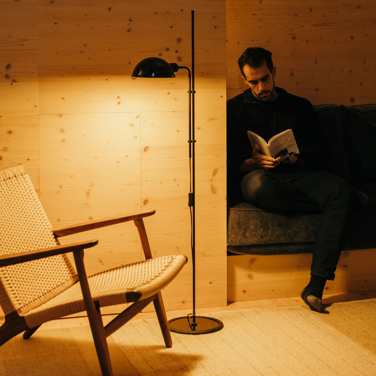 Ein mann sitzt in einem gemütlichen Raum mit Holzwänden und liest ein Buch. Neben ihm steht eine schlanke, schwarze Stehleuchte »Funiculi«, die sanftes Licht im Raum verbreitet. Ein geflochtener Holzstuhl ist ebenfalls sichtbar.