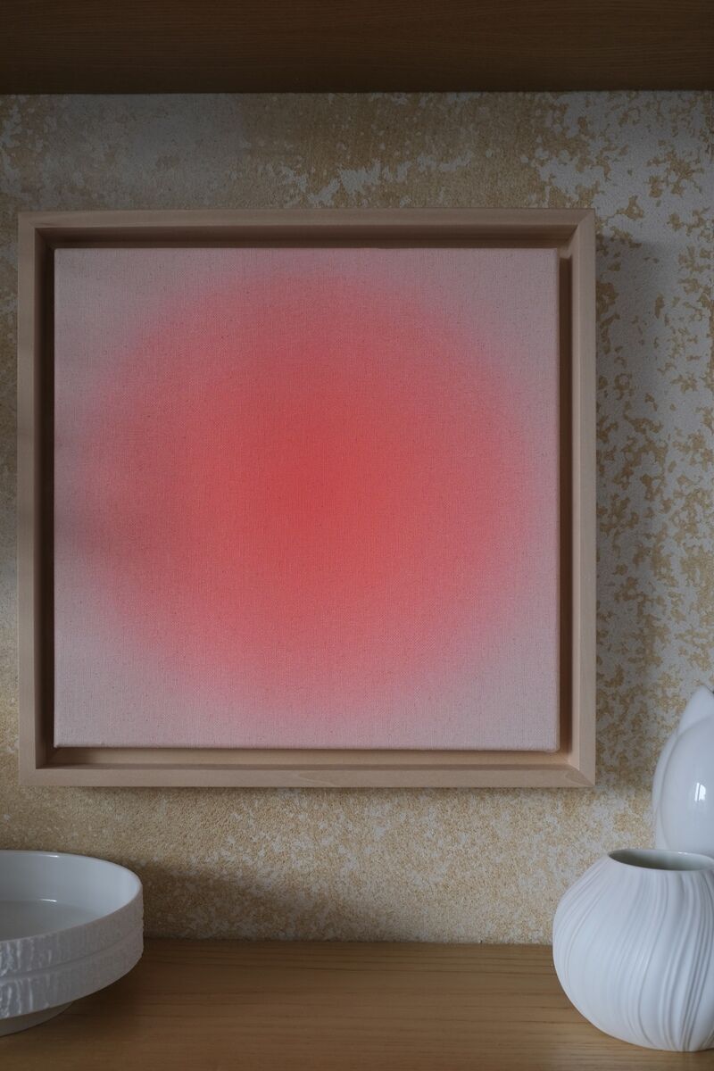 Ein quadratisches Kunstwerk mit einem roten Farbverlauf in der Mitte, gerahmt in einem naturholzfarbenen Rahmen. Es steht auf einem hölzernen Regal neben weißen Keramikdekorationen.