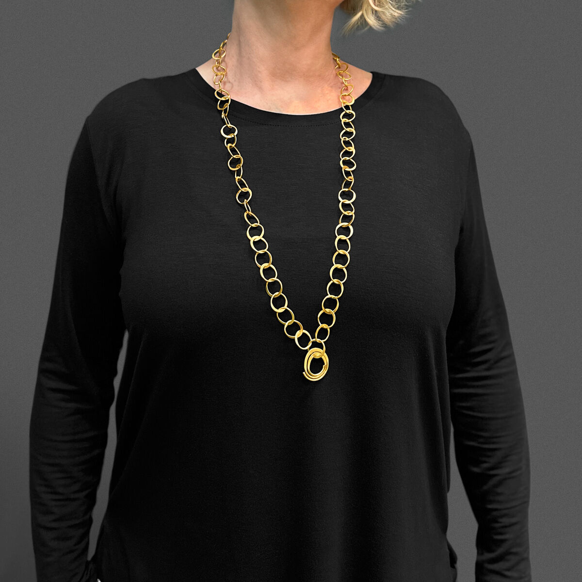 Eine Frau mit blonden Haaren und Brille trägt ein schwarzes einfaches Kleid und eine vergoldete silberne doppelte Halskette mittlerer Länge. Der Hintergrund ist grau.