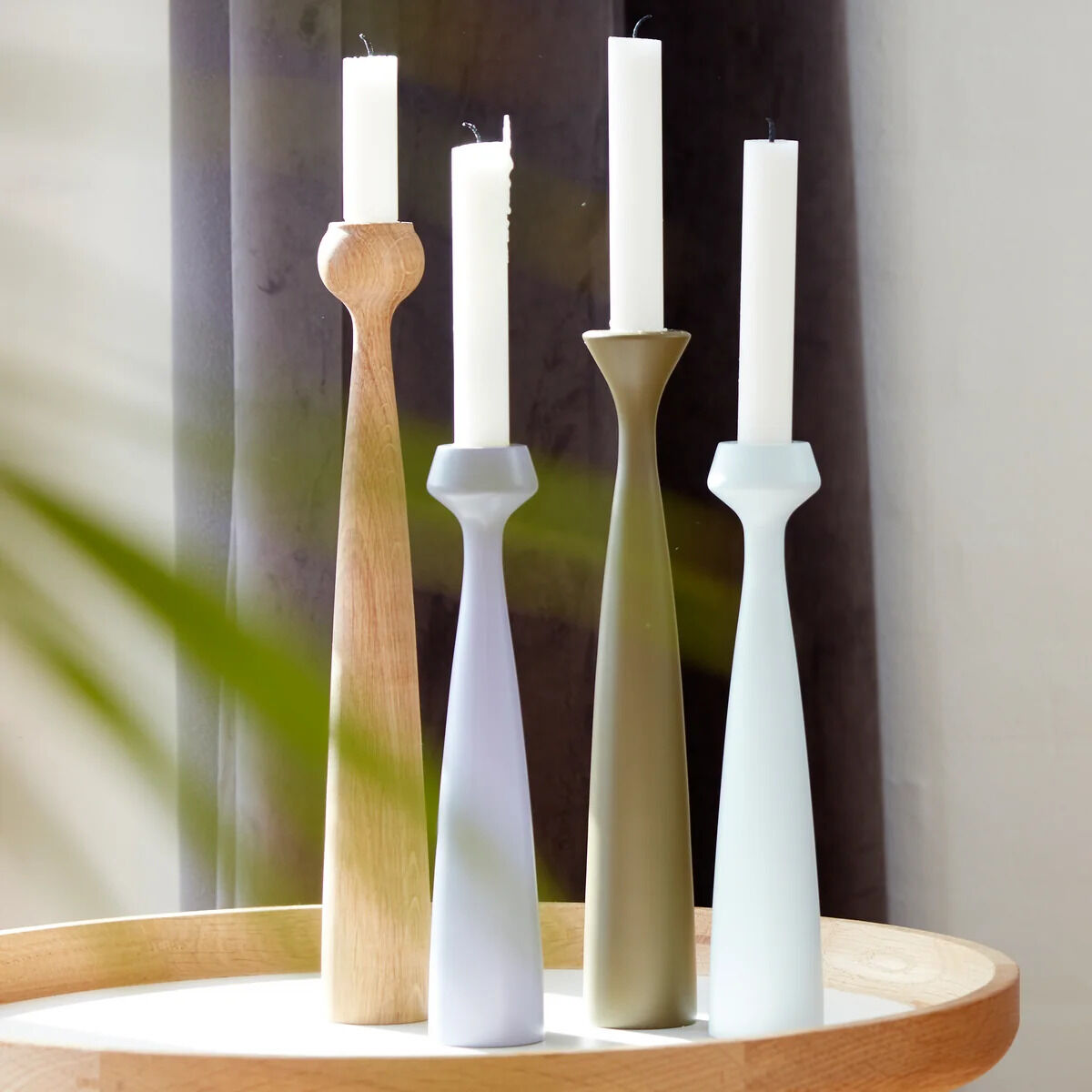 Vier elegante Kerzenhalter der Serie »Tulip« von applicata aus geölter Eiche und lackiertem Holz stehen auf einem runden hellen Tablett. Die Kerzenhalter haben eine schlanke, moderne Form, darauf weiße Stabkerzen. Licht fällt weich ins Bild.