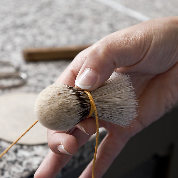 Nahaufnahme einer Hand, die einen Rasierpinsel mit feinem Dachshaar hält. Der Hintergrund zeigt eine Arbeitsfläche mit einem angefangenen Rasierset und weiteren Utensilien.