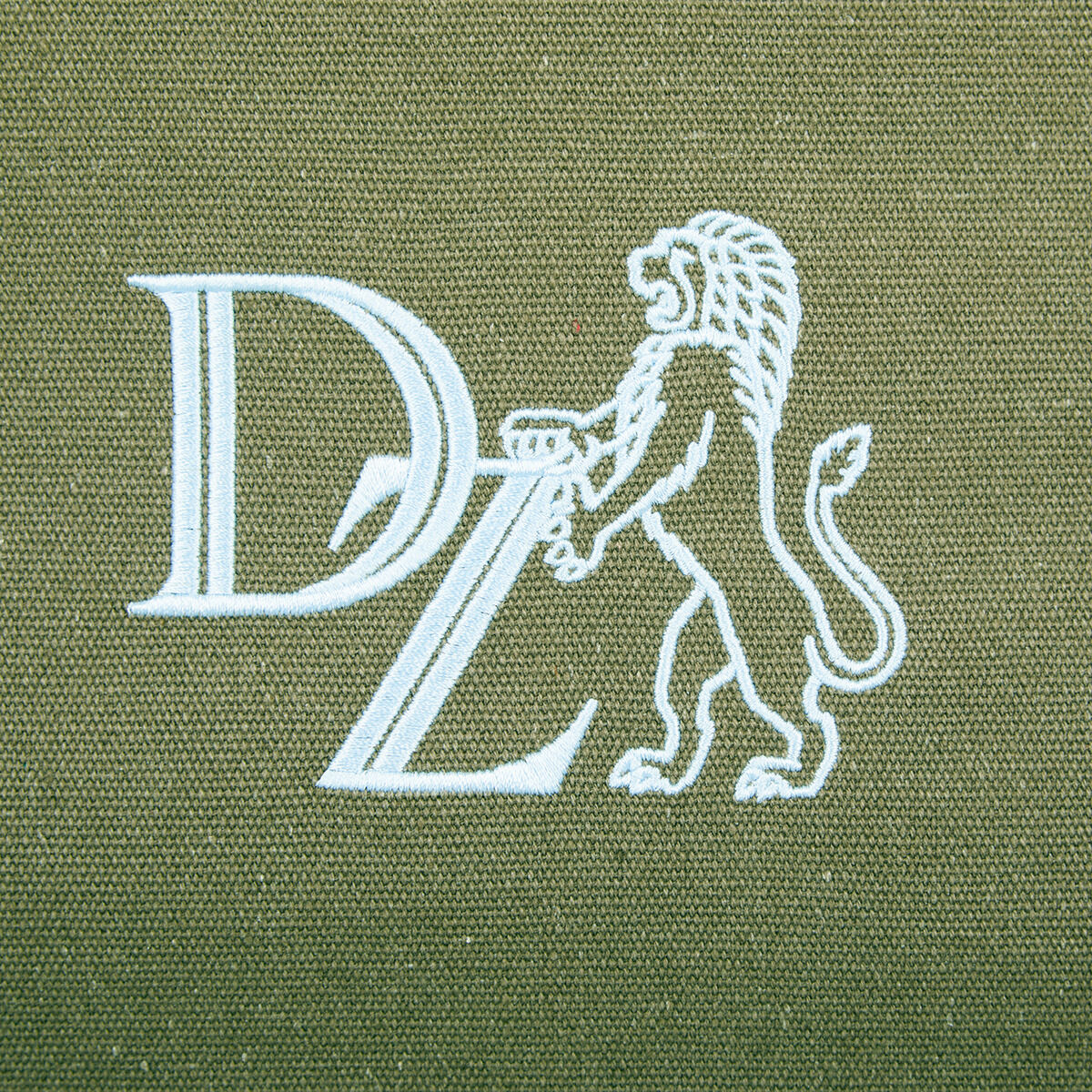 Nahe Aufnahme einer olivgrünen Canvas-Tasche. Auf der Tasche ist ein weißes Logo zu sehen, das die Buchstaben 'DZ' und ein stehender Löwe zeigt.
