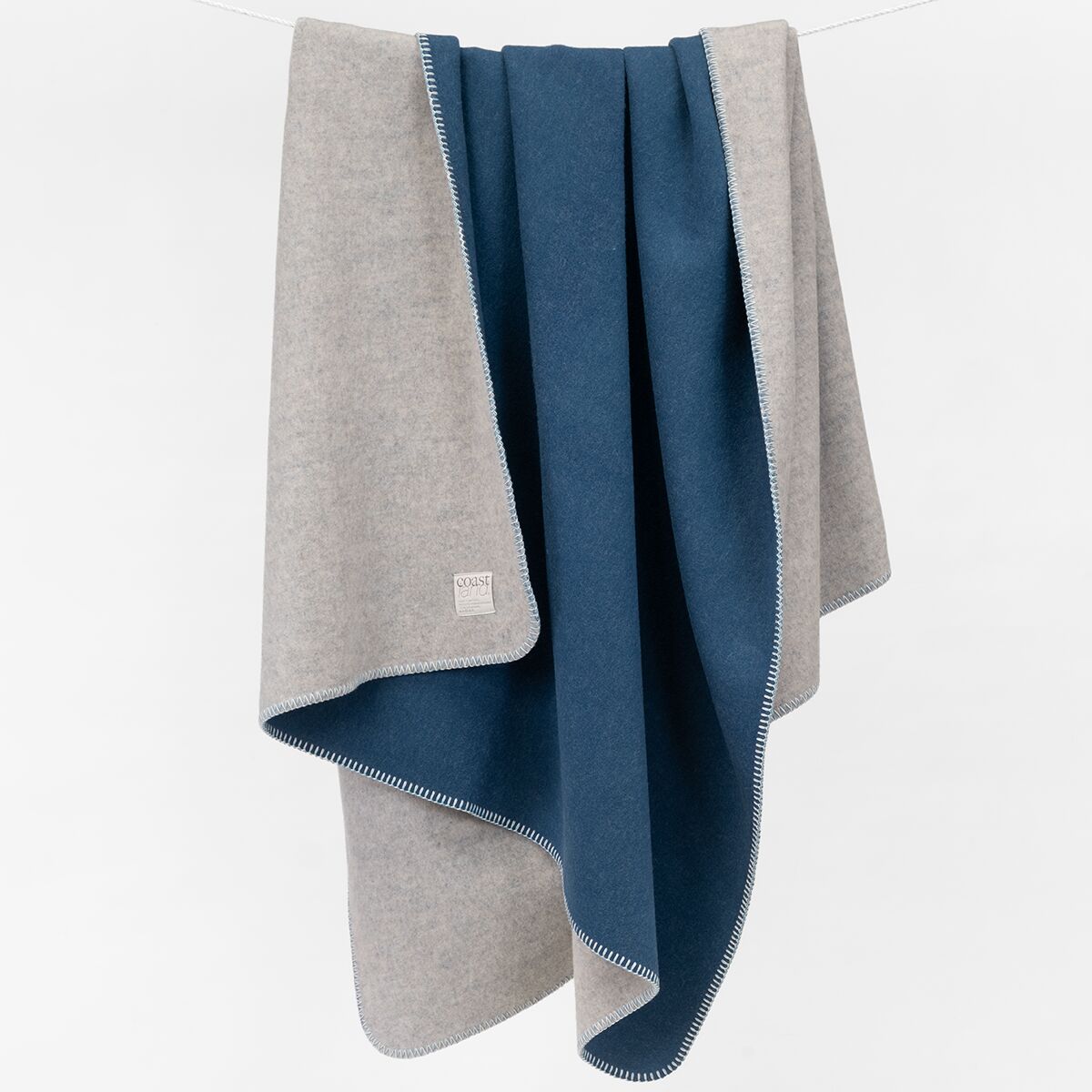 Eine zweifarbige Merinowolldecke in Blaugrau und Dunkelblau hängt ordentlich zusammengelegt. Die Decke wirkt weich und luxuriös, mit feinem Randabschluss, perfekt als Wohnaccessoire für gemütliche Stunden.
