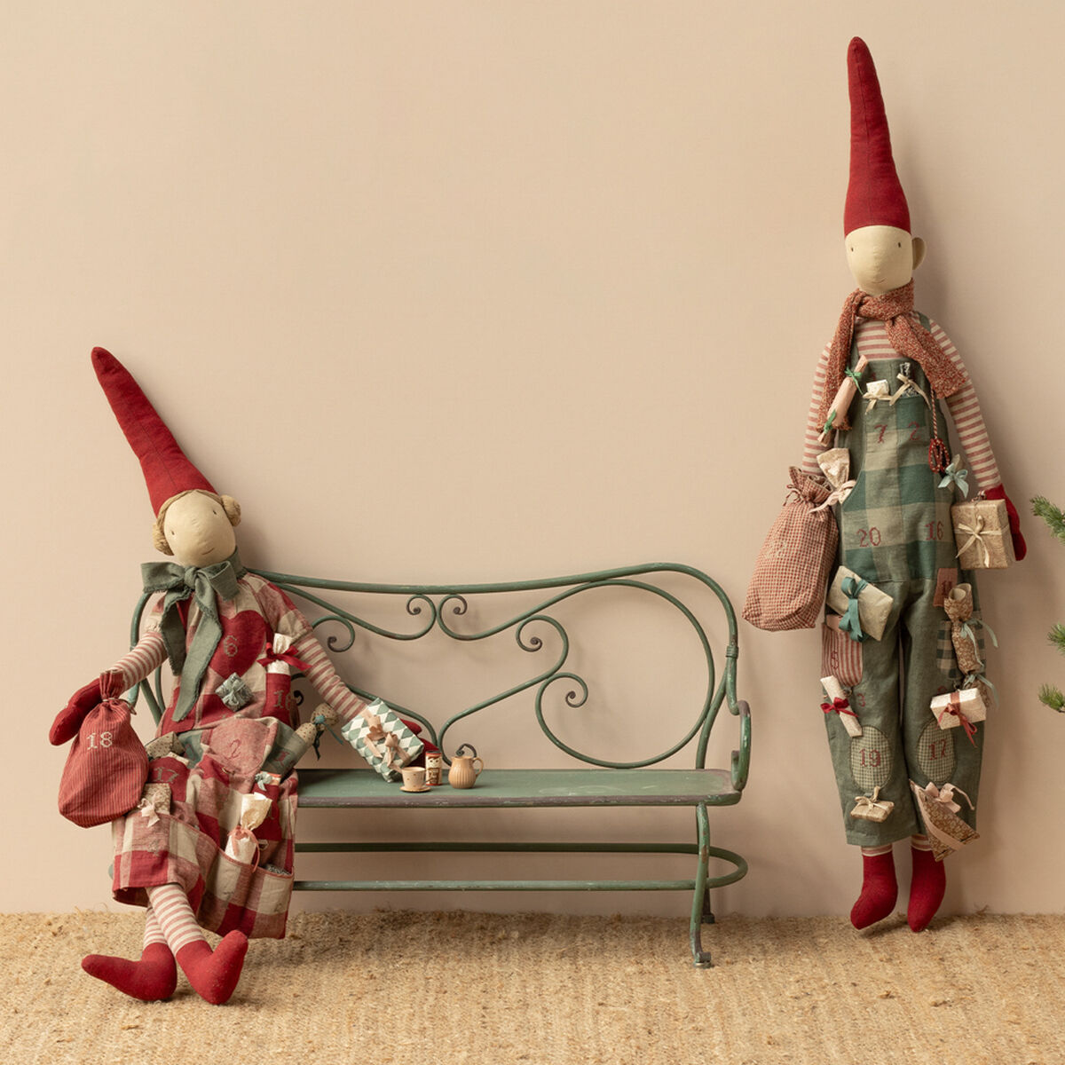Zwei Stoffpuppen in Form von Pixy Mädchen mit roten Zipfelmützen und gestreiften Kleidungsstücken stehen neben einer dekorativen Metallbank und einem kleinen Tannenbaum. Die Szene ist weihnachtlich dekoriert und die Figuren tragen kleine Geschenke sowie Adventssäckchen. Der Hintergrund ist neutral gestaltet.