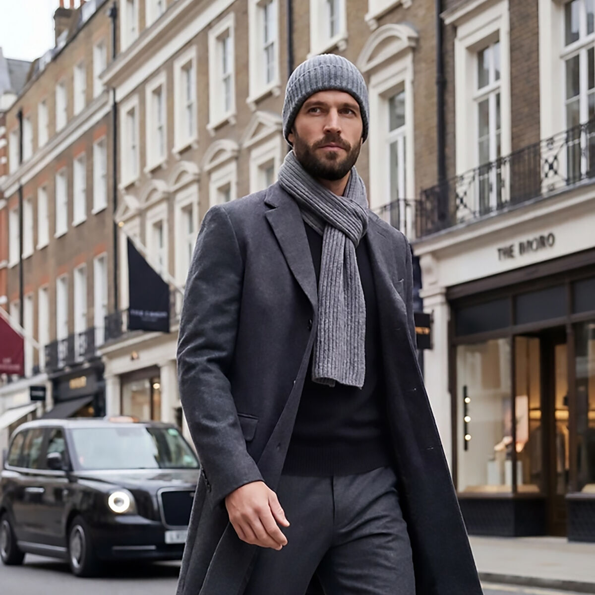 Ein stilvoll gekleideter Mann trägt einen naturbraunen Kaschmir-Schal "Dominik" von Eagle Products, grauen Mantel, graue Hose und braune Lederschuhe beim Spaziergang durch eine elegante Stadtstraße. Im Hintergrund klassische Stadthäuser und ein schwarzes Taxi.