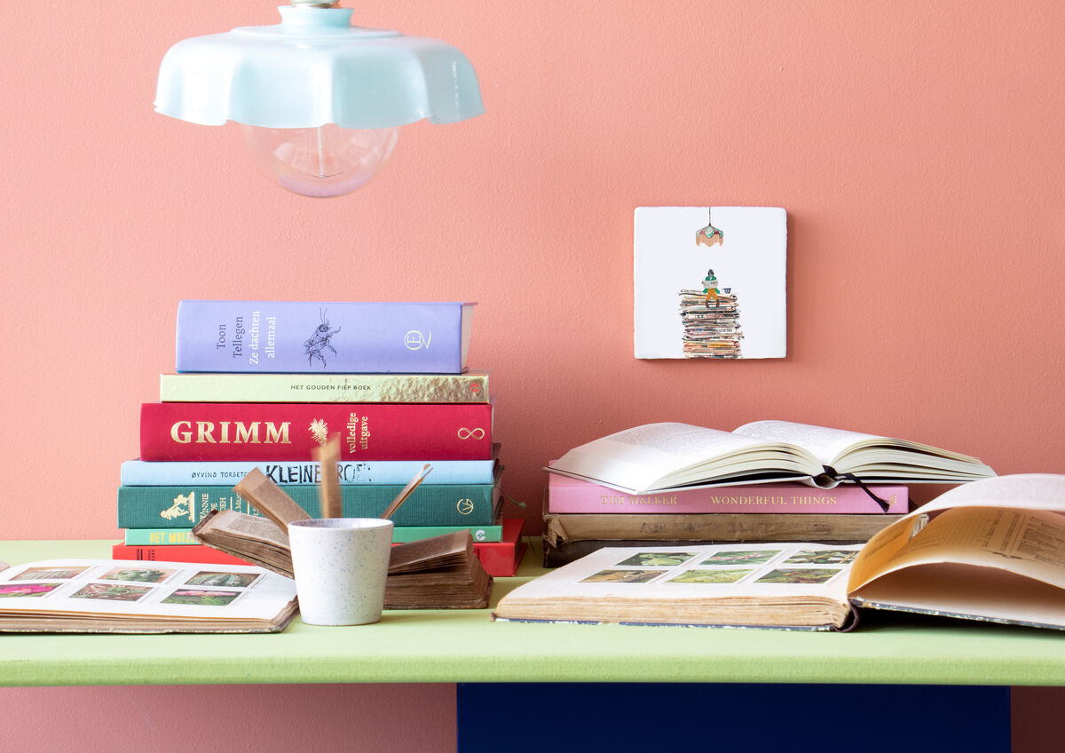 Ein Regal mit verschiedenen bunten Büchern unter einer hellen Lampe vor einer roséfarbenen Wand. Darauf steht eine Keramikfliese mit einer Buchstapel-Illustration namens »DIE ZEIT«.