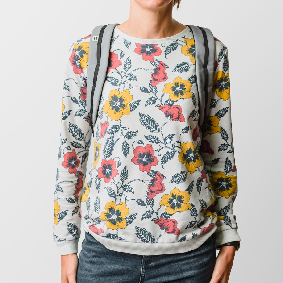 Person trägt einen floralen Pullover und einen grauen Rucksack »Wanaka«. Die Bluse ist mit gelben und roten Blumen verziert.