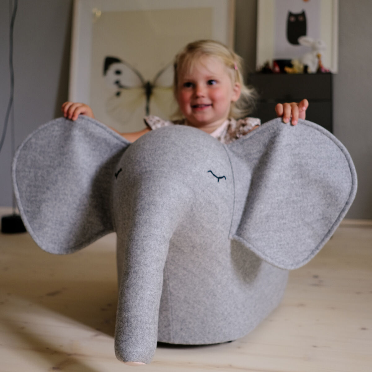 Ein kleines Kind sitzt auf einem grauen Rollelefant in einem Wohnzimmer auf einem hellen Holzboden. Im Hintergrund sind Rahmenbilder zu sehen. Das Kind lächelt und sieht glücklich aus.