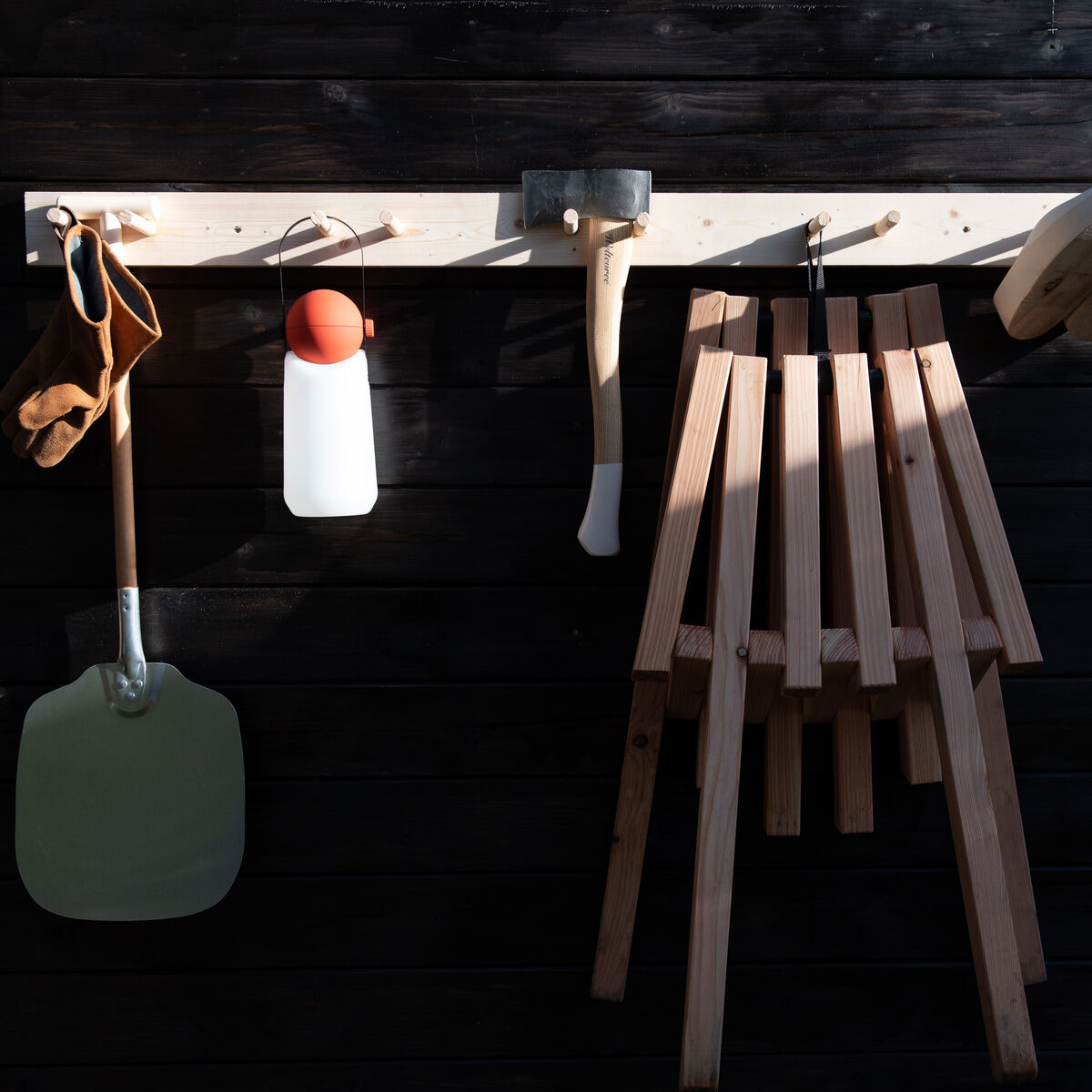 Ein Holzstuhl mit Schafsfellbezug, ideal für den Außenbereich oder Camping, der vor einer dunkel gestrichenen Holzwand hängt. Daneben sind Holzwandhaken mit Werkzeugen und Accessoires für die Gartenarbeit sichtbar.