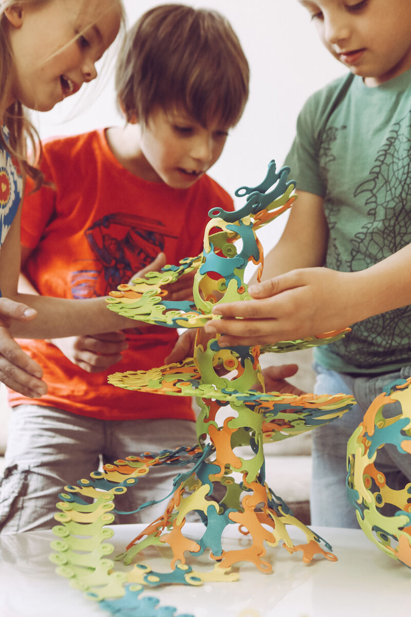 Drei Kinder spielen mit dem Konstruktionsspielzeug »Binabo« und bauen eine spiralförmige Struktur aus bunten Teilen in grün, orange, blau und gelb.