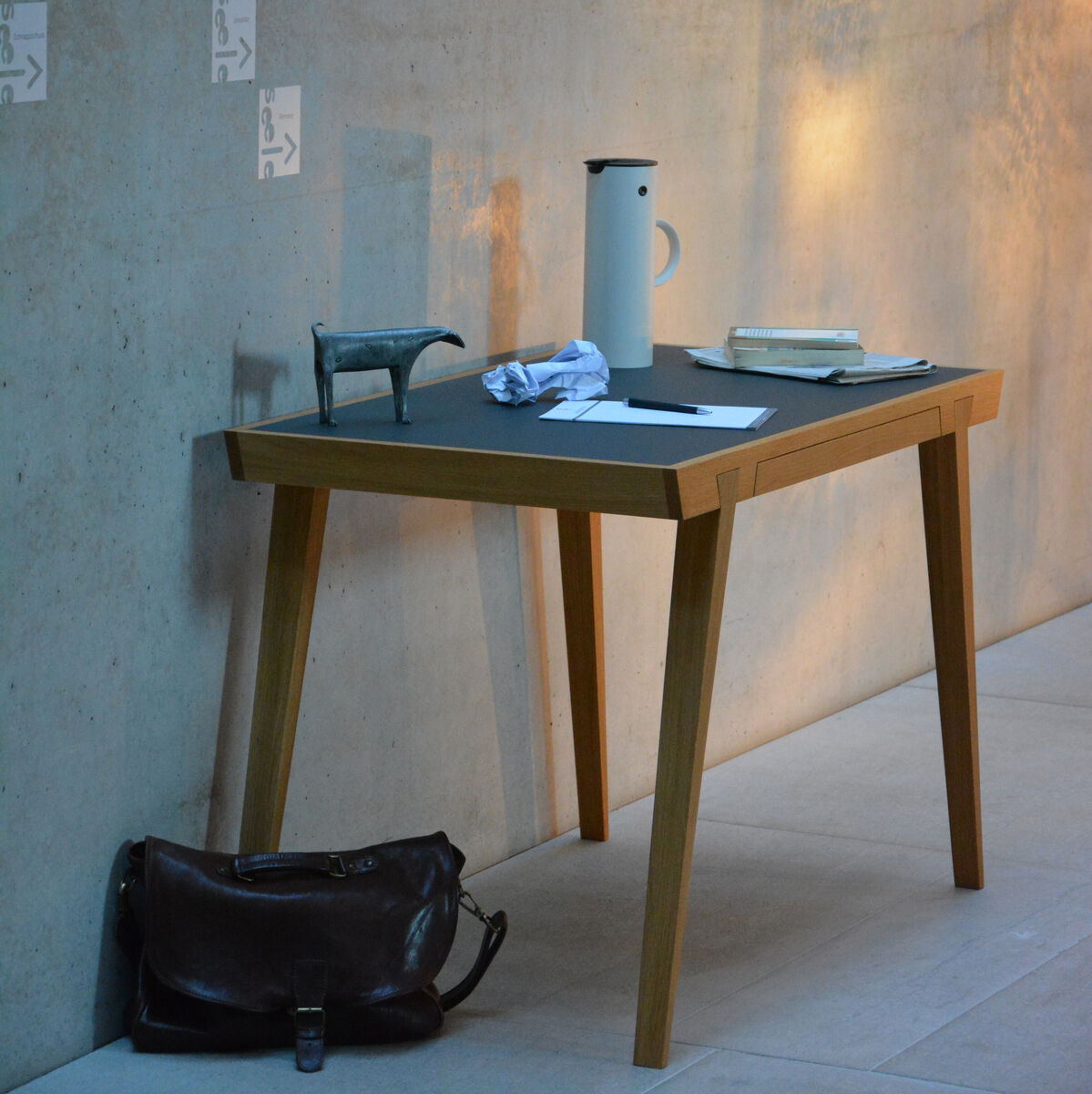 Ein moderner Schreibtisch aus Holz mit einer dunklen Tischplatte, der an einer Betonwand steht. Auf dem Tisch befinden sich eine Kanne, Bücher, Notizhefte und Dekoration. Darunter liegt eine braune Ledertasche auf dem Boden.